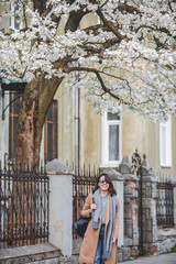 young stylish woman walking by city street in coat blooming tree on background