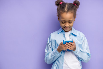 smiling cute   african child girl with mobile phone in hands, isolated over purple background. beautiful shy child chatting with friend or watching something interesting on phone