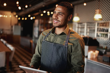 Handsome young man wearing apron holding digital tablet standing in coffee house looking away