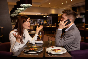 furious caucasian woman and handsome man sit quarreling, man talk on phone while his girlfriend disappointed with man's behavior
