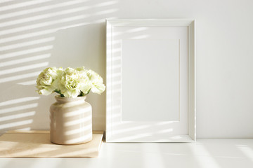 Horizontal shot of blank white photo frame for family picture and stylish ceramic vase with beautiful peony flowers, morning sunshine on wall. Interior design, coziness and decoration concept