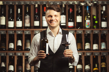 Smiling sommelier holds a bottle and glass of red wine in hands on the wine cellar background.