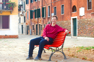 Smiling Caucasian male tourist on red bench in Italian town