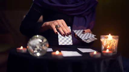 Close-up of woman fortuneteller hand with cards while sitting at table with candles in dark room