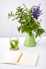 eco and organic concept - herbal tea, notebook with pencil and flowers in green jug on table