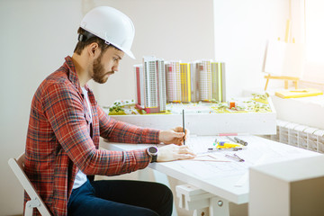 caucasian architect working in office desk, draws and takes measurements on new project, engineering and architectural concept. hard-working guy in helmet use drawings and small model of building