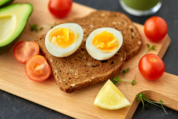 Fototapeta na wymiar food, eating and breakfast concept - toast bread with eggs, cherry tomatoes, avocado and greens on wooden cutting board