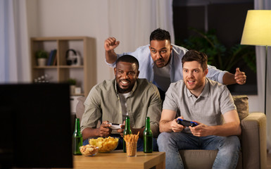 friendship, technology and leisure concept - smiling male friends with gamepads and beer playing video game at home at night