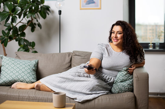 People And Leisure Concept - Happy Young Woman Lying On Sofa With Remote Control And Watching Tv At Home