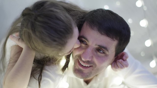 A Father in a White Shirt and a Little Girl in a White Dress are Lying on the Carpet Reading a Book Together, Joking and Fooling Around and Hugging in Christmas Decorations. New Year Family Holidays