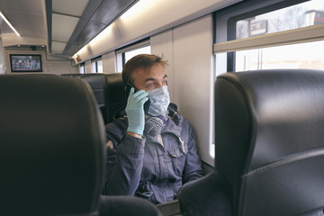 Using gadgets while disease outbreak, coronavirus covid-19 pandemic. Adult man with medical protective mask and gloves sitting inside empty train and talking on smartphone.