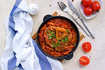 Ossobuco, beef shank stew on the bone with carrots, tomatoes and celery in a serving cast-iron frying pan on a light concrete background. Main dishes. Italian food.