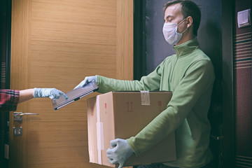 Courier in protective mask delivers parcel to client, client in medical gloves signs on tablet....