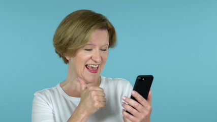 Fototapeta premium The Old Woman Excited for Success while Using Smartphone Isolated on Blue Background