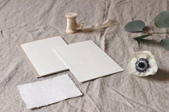 Wedding stationery mock-up scene. Blank greeting cards on linen tablecloth background with white anemone flower, eucalyptus tree branches and silk ribbon. Feminine still life composition. High angle