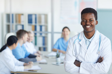 Waist up portrait of young African-American doctor smiling at camera while standing with arms crossed against medical conference background, copy space