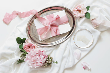 Feminine wedding, birthday mock-up scene. Blank paper greeting cards, pink roses, silk ribbons, peony flowers on silver plate, White table background. Light, shadow play. Flat lay, top view