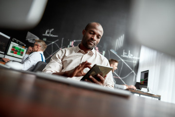 The best financial state of mind. Focused african male trader sitting by desk and studying analytical reports using tablet pc in the office. His colleagues are working in the background