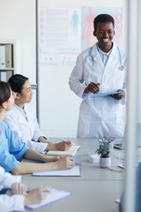 Portrait of young African-American doctor giving speech while standing at table during medical council or conference in clinic, copy space