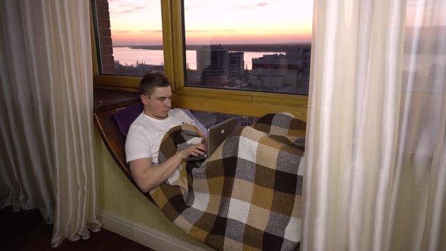 A young man is typing on a laptop. A man lies on a window sill by the window with a laptop in his hands. Outside the evening
