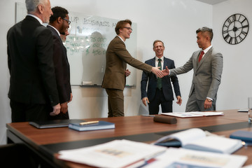 Good deal. Caucasian and Asian businessmen in classic wear shaking hands after signing the agreement while standing with their team in the modern office