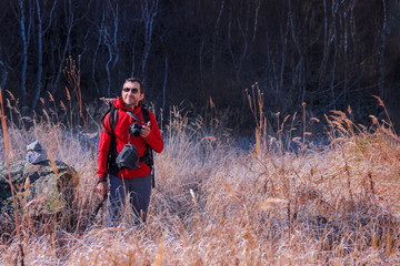Naklejka na ściany i meble A smiling middle-aged man in a red jacket and glasses with a camera watches birds in the tall grass in autumn.