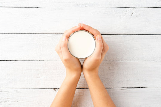 Overhead Shot Of Woman’s Hands Holding Glass Of Milk On White Wooden Table