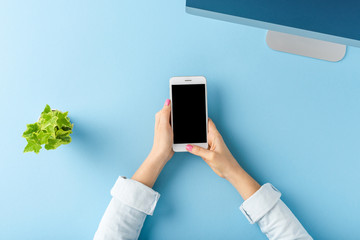 Woman’s hands using smart phone with empty screen on blue table. Office desktop. Flat lay