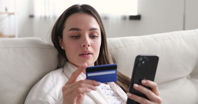 Happy young woman holding credit card using instant mobile payments at home. Smiling beautiful girl customer shopper making purchase on smart phone receiving cashback concept. E-banking app service