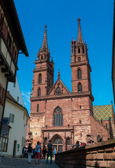 Naklejka na ściany i meble Great full view of the famous landmark Basel Minster. Tourists are taking photos of the beautiful church with its red sandstone walls, colourful roof tiles and impressive twin towers. 