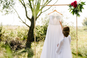 the bride in a white coat and with a veil touches a white dress that hangs on a white bar in nature