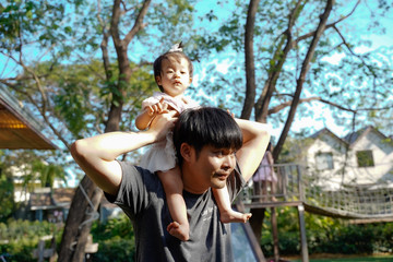 Fototapeta premium Asian daughter riding her father's neck and hold father hand in sunny day, Dad and baby girl playing together walking in the garden in playground park. 