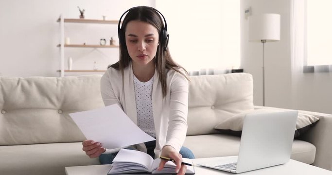 Young woman entrepreneur wearing wireless headset making conference business call working from home sitting on sofa. Female distant online teacher doing video chat communicating by webcam on laptop.