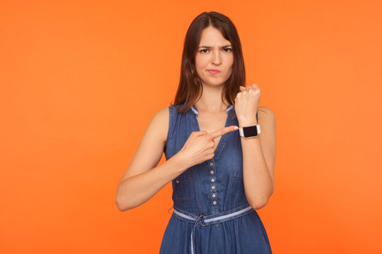 Worried brunette woman in denim dress being punctual, looking anxious and impatient about late hour, delay or deadline, pointing to wristwatch, showing clock. studio shot isolated on orange background