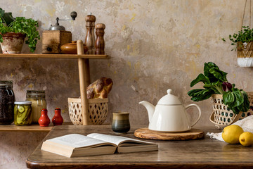 Stylish composition of kitchen interior with family table, vegetables, tea pot, dessert, shelf with food supplies, plants and kitchen accessories in wabi sabi concept of home decor. 