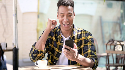 The Young African Man Celebrating Success on Smartphone, Outdoor Cafe