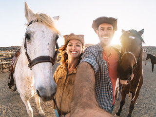 Happy couple taking selfie with horses inside stable - Young farmers having fun with animals in...