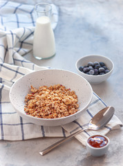 Breakfast, hand pours milk from a jug into muesli in a deep dish. Next to blueberries and jam on a blue-gray concrete background