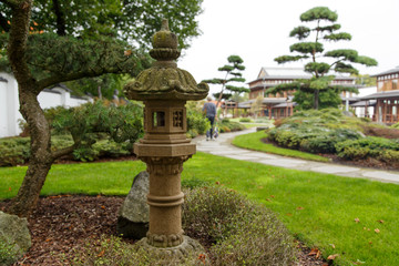 asian antique stone lantern with path on which a woman walks through park with asian topiaries and solitary shrubs
