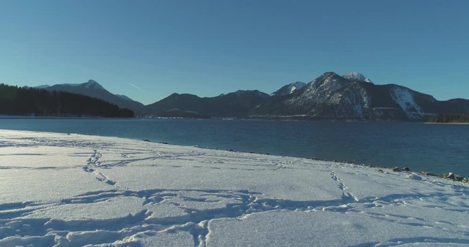 4K Drone aerial footage of the famous, scenic and idyllic Bavarian lake Walchensee in southern Germany in the alps. The shore is covered in fresh white snow with some footsteps on a sunny afternoon.