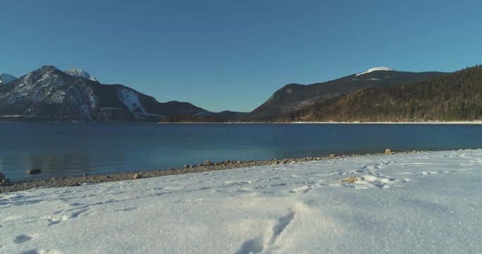 4K Aerial Drone footage of the famous, scenic and idyllic Bavarian lake Walchensee in southern Germany in the alps. The shore is covered in fresh white snow with some footsteps on a sunny afternoon.
