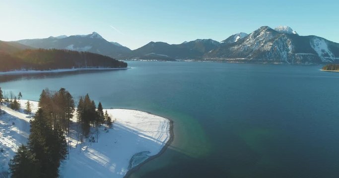 4K Aerial Drone footage of the famous, scenic and idyllic Bavarian lake Walchensee in southern Germany in the alps. The shore is covered in fresh white snow with pine trees on a sunny afternoon.