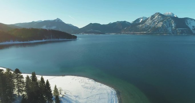 4K UHD Aerial Drone footage of the famous, scenic and idyllic Bavarian lake Walchensee in southern Germany in the alps. The shore is covered in fresh white snow with mountains on a sunny afternoon.