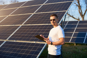 Young stylish guy in sunglasses makes an entry in folder against the background of rows solar panels under clear blue sky. Man with watch in white T-shirt and jeans is smiling while looking at camera