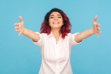 Fototapeta premium Come here, free hugs! Portrait of hipster woman with fancy red hair in shirt smiling genuinely and stretching arms to camera, welcoming ready to embrace. indoor studio shot isolated on blue background