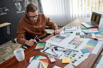 Creative worker. Concentrated male ux designer in a wheelchair choosing color samples for mobile app while working at his creative workplace in the modern office