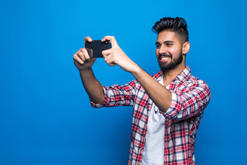 Young happy bearded indian man take photo using mobile phone posing isolated over blue wall...