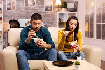 In modern cozy living room couple is enjoying takeaway noodles while watching TV
