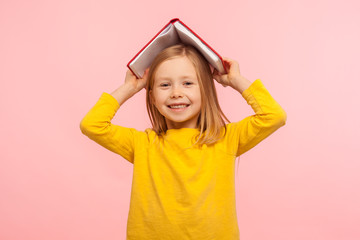 Portrait of happy lazy little girl covering head with book and smiling to camera, disobedient child...