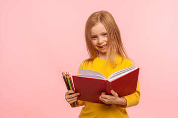 Education, elementary school. Portrait of adorable little girl smiling to camera, holding big book and pencils, development of creative child abilities. indoor studio shot isolated on pink background